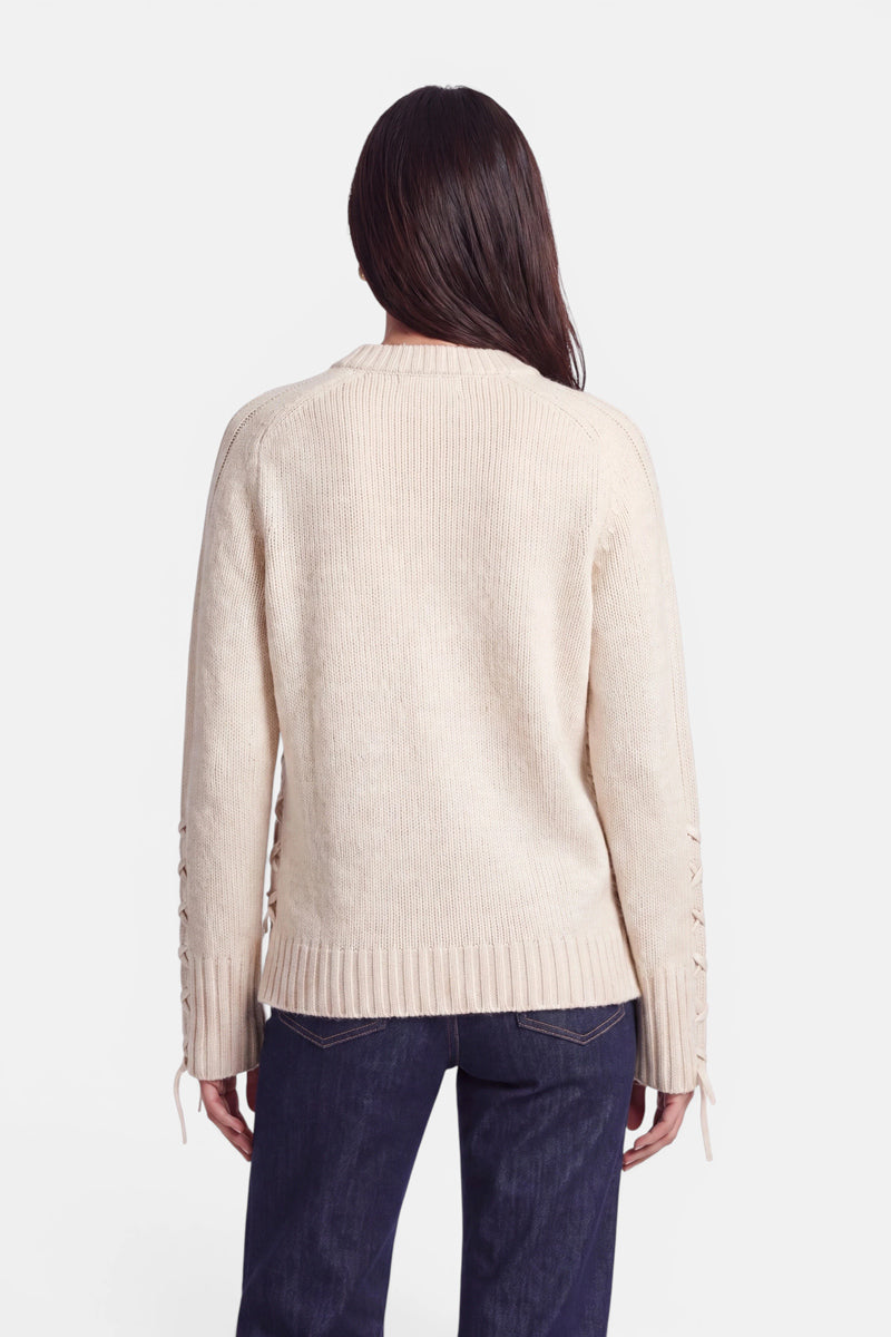 Eine Person mit langen, glatten, dunklen Haaren trägt einen hellbeigen Pullover aus Alpaka-Wolle mit geripptem Saum und Bündchen sowie dunkelblaue Jeans. Das Bild zeigt die Person von hinten vor einem schlichten, weißen Hintergrund. Der Pullover weist Schnürdetails an den Ärmeln auf.