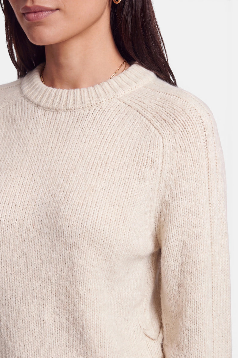 Eine Frau, die von den Schultern aufwärts zu sehen ist und einen cremefarbenen Pullover aus Alpaka-Wolle mit geripptem Rundhalsausschnitt trägt. Ihr dunkles Haar ist glatt und offen getragen, und sie trägt einen kleinen goldenen Creolen-Ohrring. Der Hintergrund ist schlicht weiß.