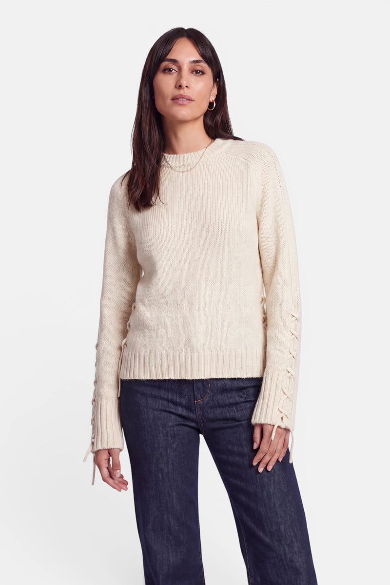 Eine Frau mit langen dunklen Haaren trägt einen cremefarbenen Pullover aus Alpaka-Wolle mit Schnürdetails an den Ärmeln und dunkelblaue Jeans. Sie steht mit dem Blick nach vorne vor einem schlichten weißen Hintergrund, mit neutralem Gesichtsausdruck und entspannter Körperhaltung.