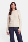 Eine Frau mit langen dunklen Haaren trägt einen cremefarbenen Pullover aus Alpaka-Wolle mit Schnürdetails an den Ärmeln und dunkelblaue Jeans. Sie steht mit dem Blick nach vorne vor einem schlichten weißen Hintergrund, mit neutralem Gesichtsausdruck und entspannter Körperhaltung.