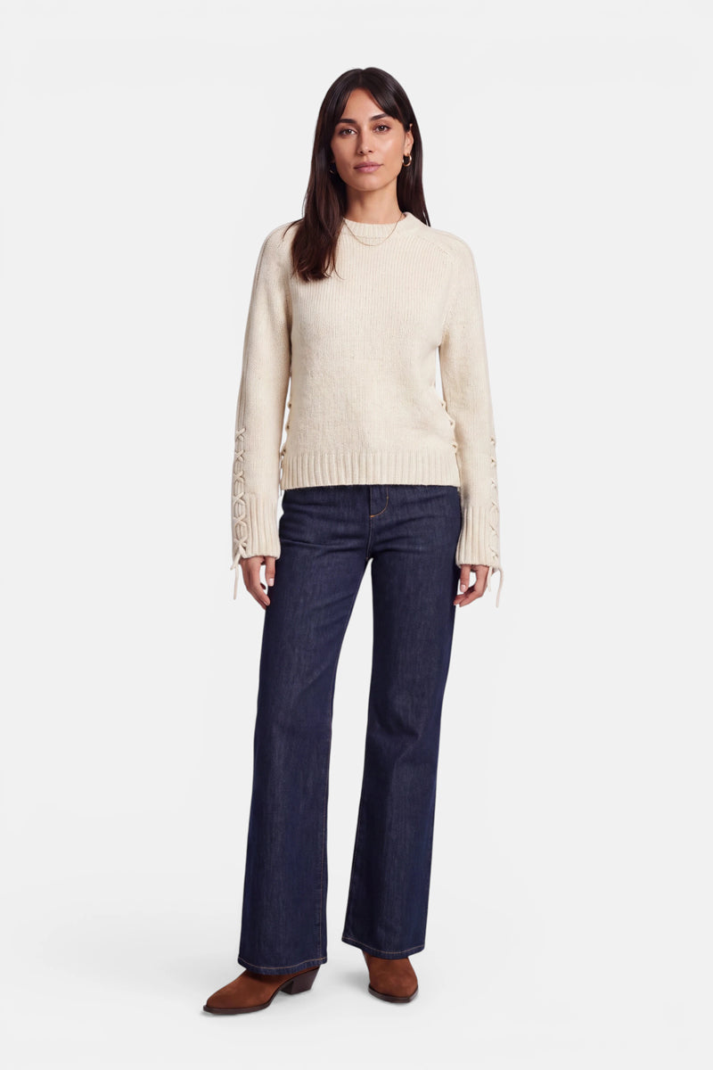 Eine Frau steht mit dem Blick nach vorne und trägt einen cremefarbenen Pullover aus Alpaka-Wolle mit eleganten Schnürdetails an den Ärmeln, dunkelblaue Jeans mit weitem Bein und braune Schuhe. Sie hat glattes, dunkles Haar und einen neutralen Gesichtsausdruck. Der Hintergrund ist schlicht weiß.