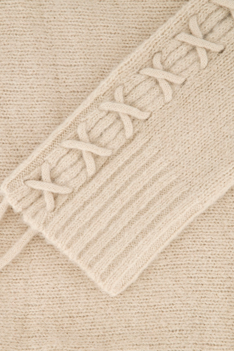 Nahaufnahme eines beigen Pullovers aus Alpaka-Wolle, die einen Detailausschnitt mit Schnürdetails in kreuz und quer verlaufenden Schnürbändern und einer gerippten Struktur zeigt. Das Kreuzmuster verläuft diagonal über das Oberteil und bildet einen Kontrast zum glatteren, einfarbigen Strickhintergrund.