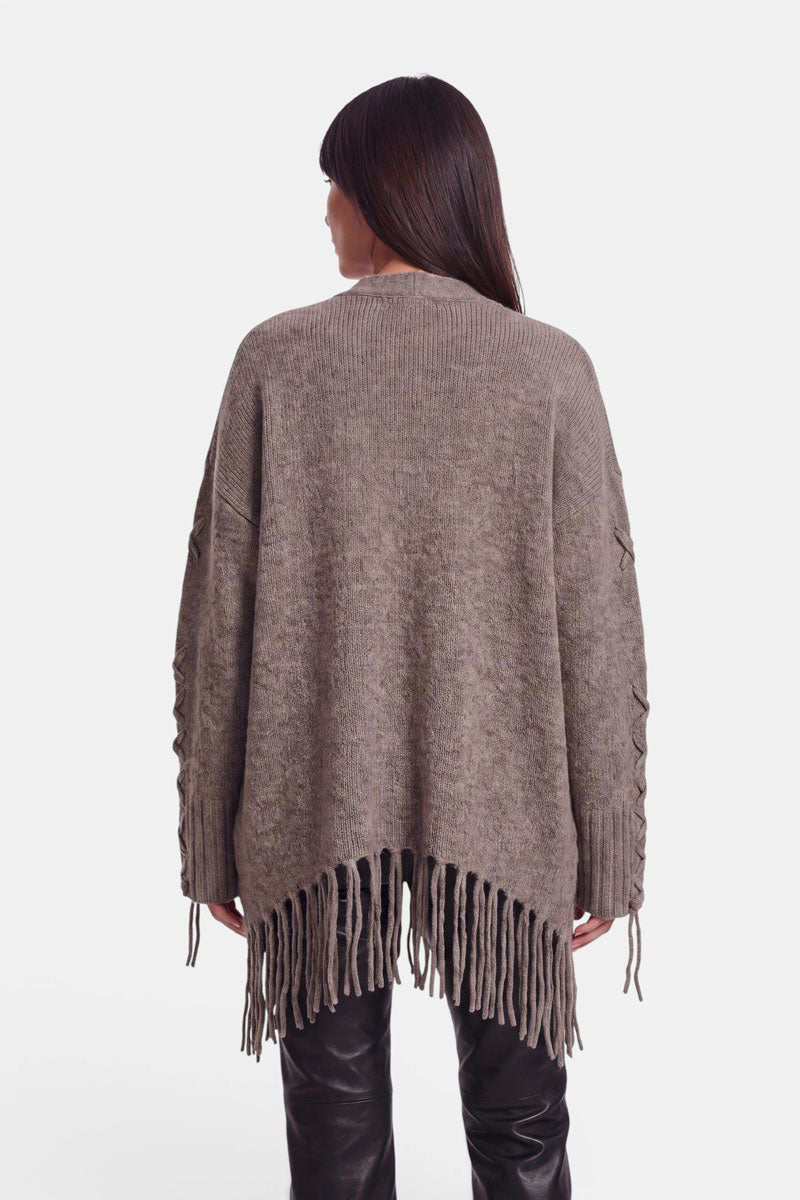 Eine Person mit langen, dunklen Haaren ist von der Seite zu sehen. Sie trägt eine locker sitzende, taupefarbene Strickjacke aus einem Alpaka-Woll-Mix mit langen Fransen am Saum und Schnürdetails an den Ärmeln, dazu eine schwarze Lederhose. Der Hintergrund ist schlicht weiß.