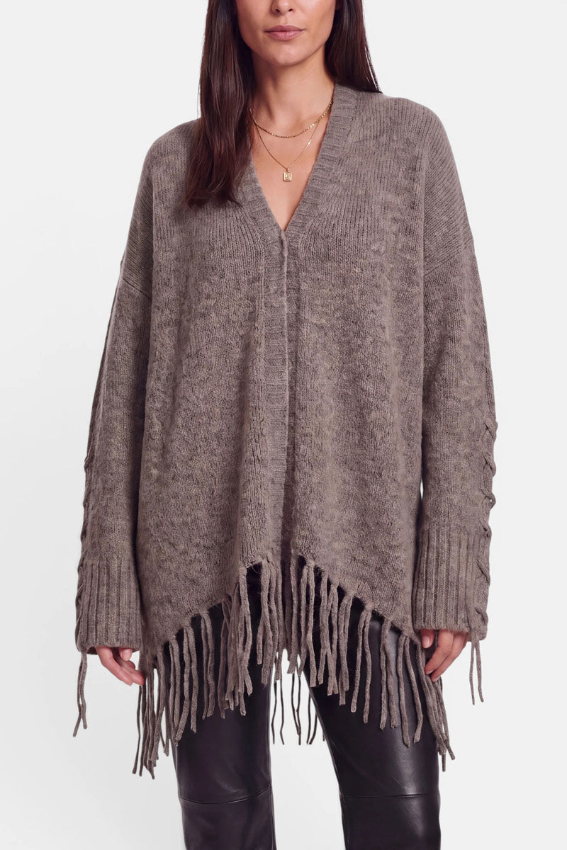 Eine Frau trägt eine locker sitzende, taupefarbene Strickjacke aus einem Alpaka-Woll-Mix mit V-Ausschnitt, langen Ärmeln mit Schnürdetails und langen Fransen am Saum. Dazu trägt sie eine schwarze Lederhose und mehrere goldene Halsketten übereinander. Ihr Haar ist glatt und offen getragen.