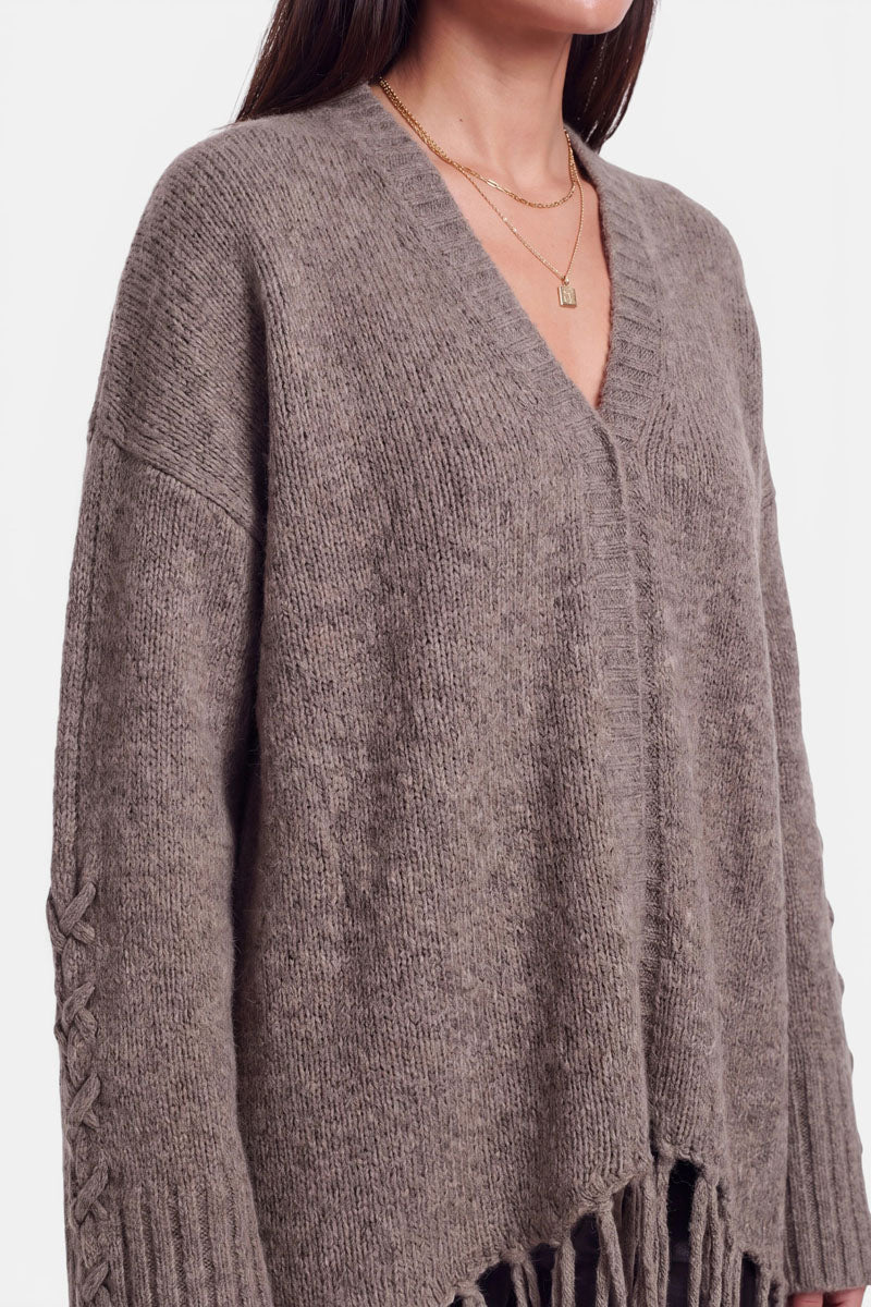 Eine Frau trägt eine locker sitzende, taupefarbene Strickjacke mit V-Ausschnitt und weiten Ärmeln, die mit einem geflochtenen Detail verziert sind. Die Strickjacke aus einer Alpaka-Woll-Mischung hat einen Saum mit Fransen. Sie hat glattes, dunkelbraunes Haar und trägt zwei goldene Halsketten. Das Foto zeigt sie von der Schulter bis zur Hüfte.