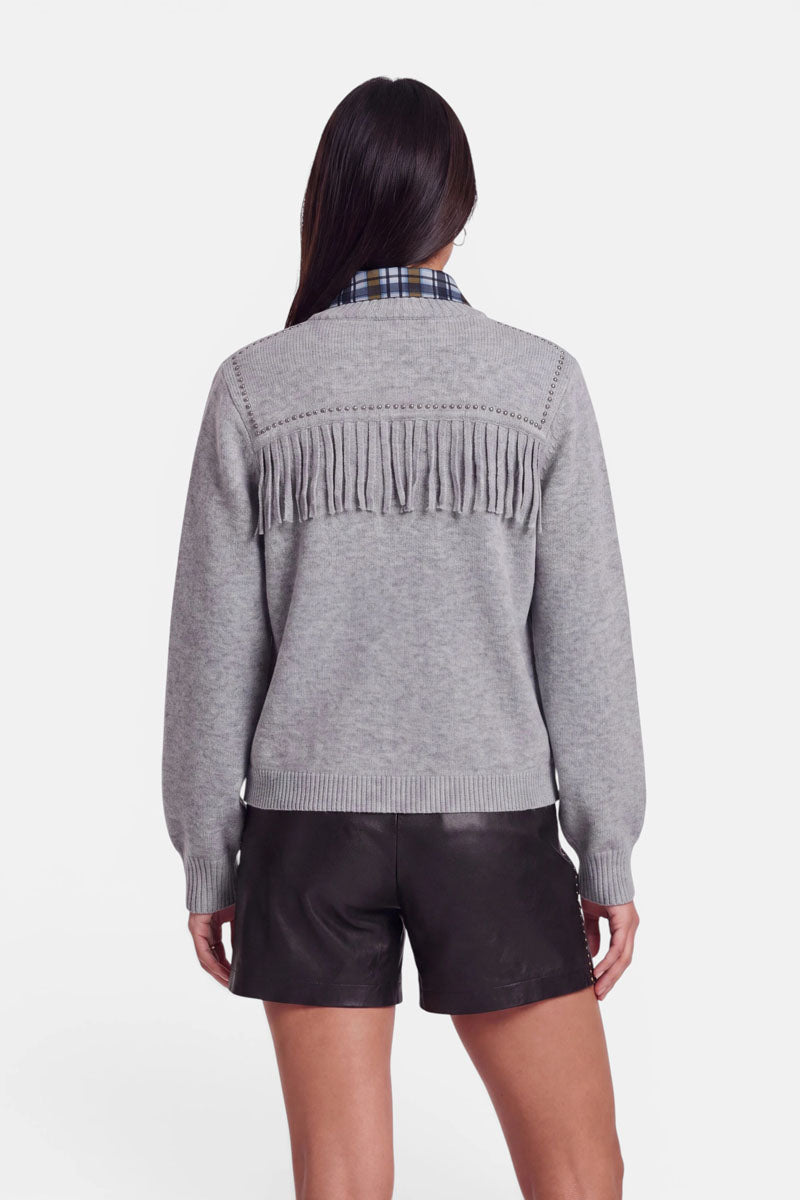Eine Frau steht mit dem Rücken zur Kamera und trägt eine graue Strickjacke aus Merinowolle mit Fransen am hinteren Passe, unter der ein blauer Karukragen hervorschaut, sowie schwarze Ledershorts. Ihr langes, dunkles Haar fällt ihr offen über die Schultern. Der Hintergrund ist schlicht weiß.