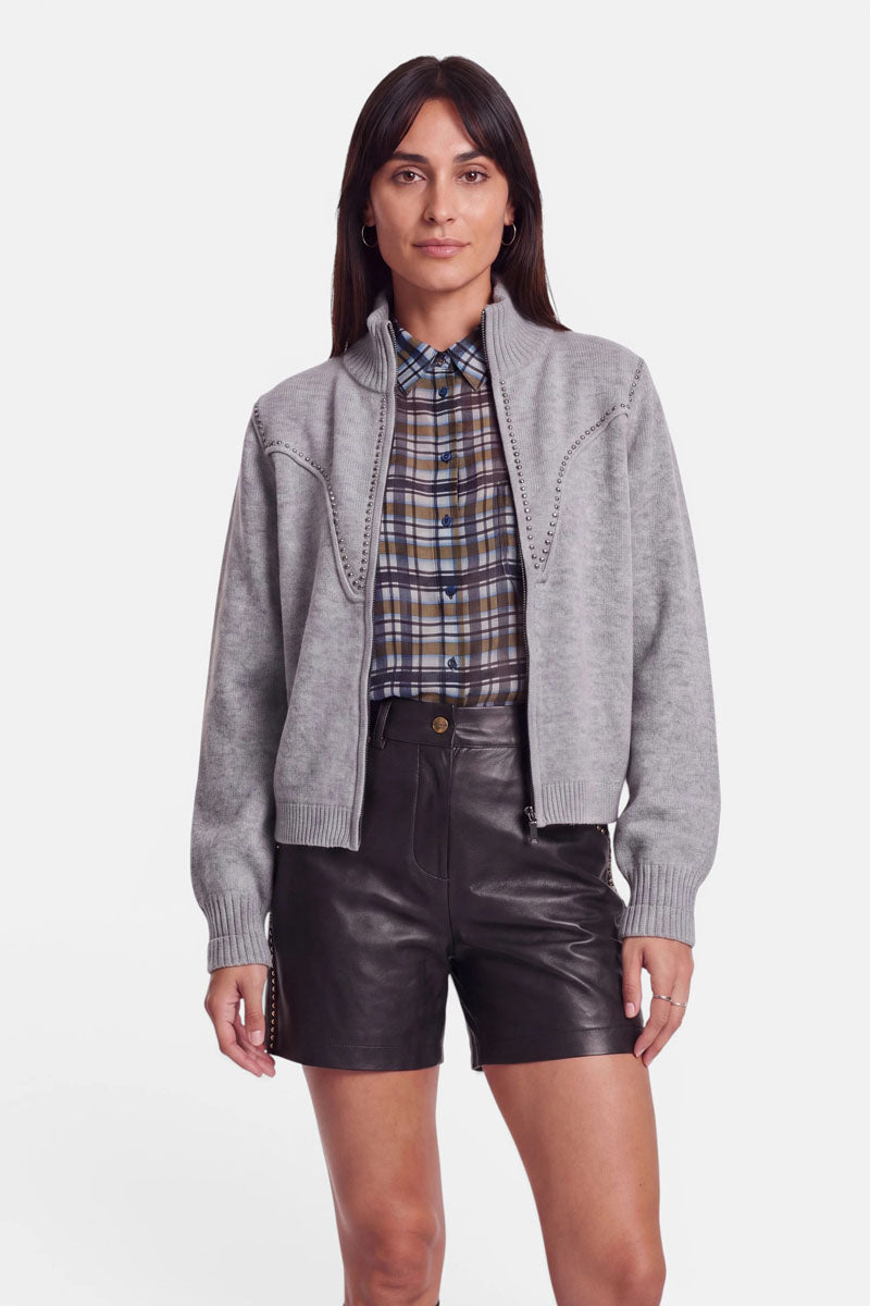 Eine Frau steht mit dem Blick nach vorne und trägt einen grauen Cardigan mit Zwei-Wege-Reißverschluss über einem karierten Hemd und schwarzen Ledershorts mit hoher Taille. Sie hat glattes, dunkles Haar, ist natürlich geschminkt und hat einen neutralen Gesichtsausdruck. Der Hintergrund ist schlicht weiß.