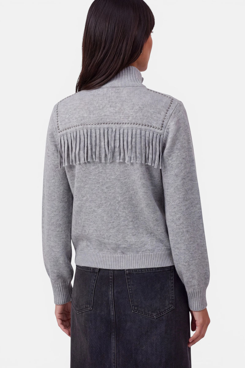 Eine Frau, die mit dem Rücken zur Kamera steht und eine hellgraue Strickjacke mit Fransen und strukturierten Details im oberen Rückenbereich trägt. Die aus weicher Merinowolle gefertigte Strickjacke hat einen gerippten Kragen, gerippte Ärmelbündchen und einen gerippten Saum. Außerdem trägt sie einen dunkelgrauen, hoch taillierten, knielangen Jeansrock. Der Hintergrund ist einfarbig weiß.