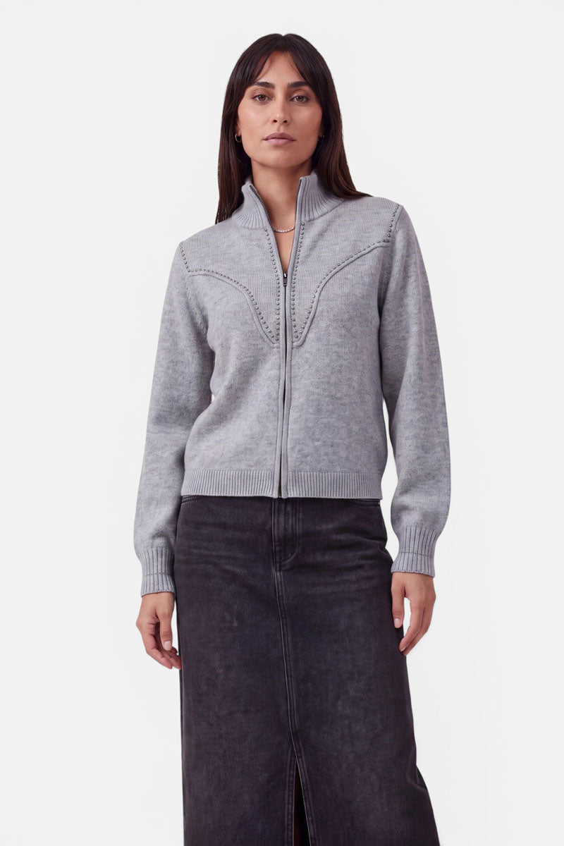 Eine Frau mit glattem, dunklem Haar trägt eine hellgraue Strickjacke aus Merinowolle mit Reißverschluss und gerippten Bündchen sowie einen langen, dunkelgrauen Rock. Sie steht mit dem Gesicht nach vorne vor einem schlichten weißen Hintergrund und blickt mit neutralem Gesichtsausdruck direkt in die Kamera.