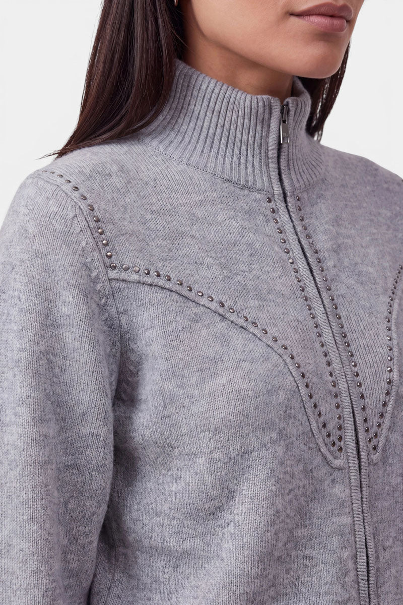 Eine Frau trägt eine hellgraue, gerippte Strickjacke mit Reißverschluss und hohem Kragen. Der aus weicher Merinowolle gefertigte Pullover ist mit kleinen Metallnieten verziert, die über Brust und Schultern ein geschwungenes, dekoratives Muster bilden. Er ist mit einem praktischen Zwei-Wege-Reißverschluss versehen. Zu sehen sind nur ihr unterer Gesichtsbereich und ihr oberer Oberkörper.