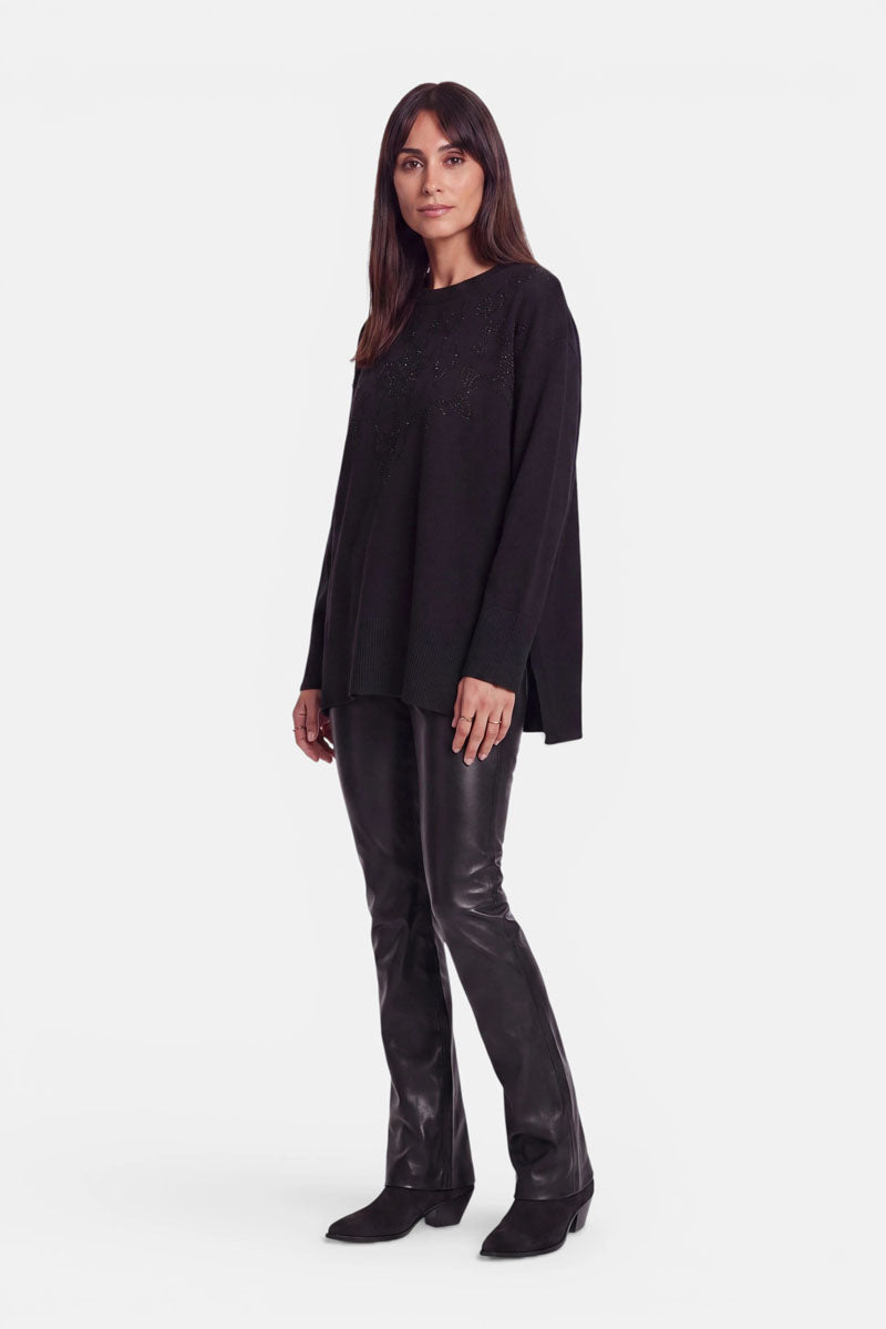 Eine Frau steht mit dem Blick nach vorne und trägt einen locker sitzenden Pullover aus Merino und Kaschmir sowie eine schwarze Lederhose. Sie hat langes, glattes, dunkles Haar und schwarze Schuhe. Der Hintergrund ist schlicht weiß, Strassverzierungen setzen dezente Akzente auf ihrem Outfit, und ihr Gesichtsausdruck ist neutral.