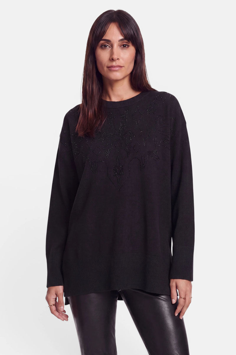 Eine Frau mit glattem, dunkelbraunem Haar, die einen locker sitzenden schwarzen Pullover aus Merino-Kaschmir mit dezenter Strassverzierung und eine schwarze Lederhose trägt, steht mit dem Blick nach vorne vor einem schlichten weißen Hintergrund.