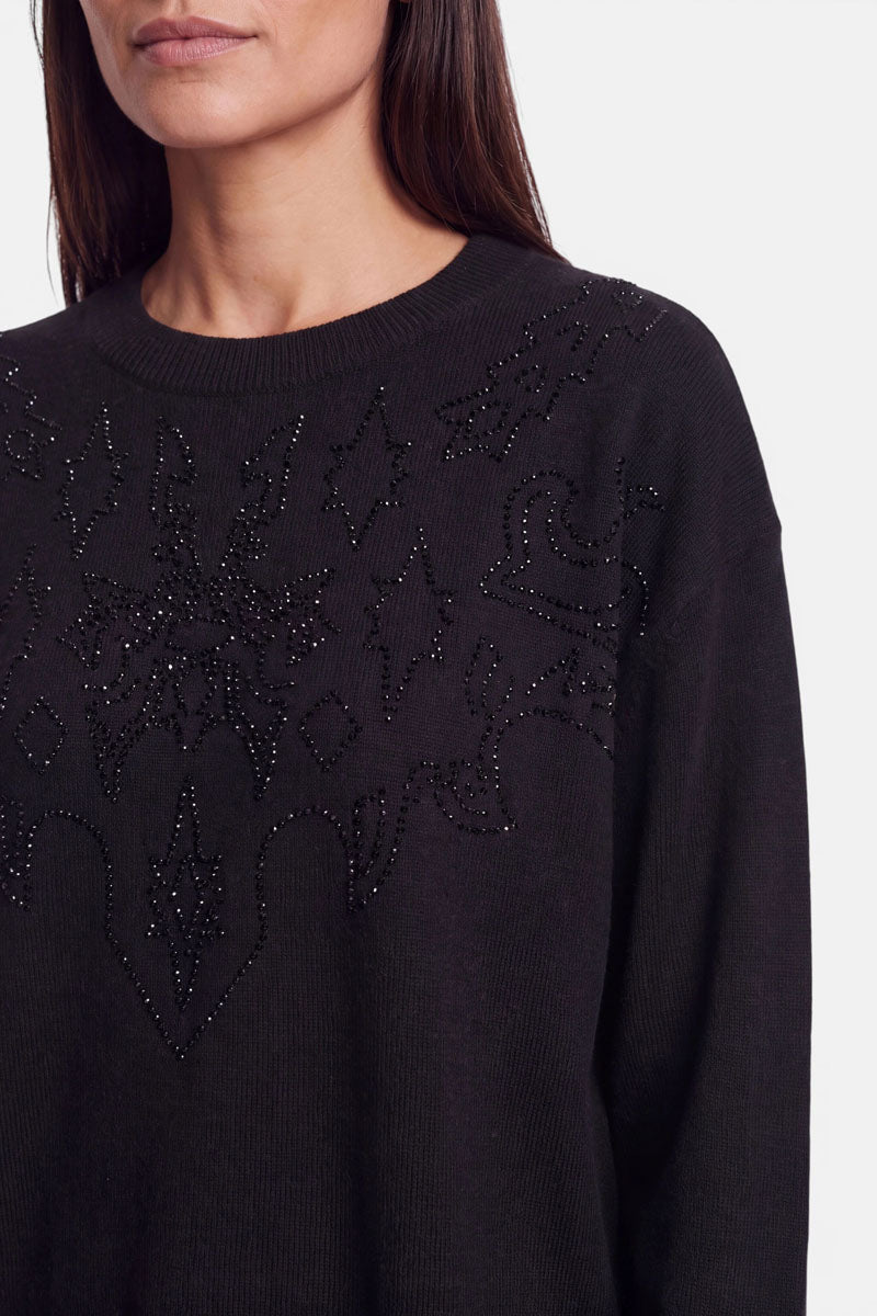 Eine Frau mit glattem, dunklem Haar trägt einen schwarzen Pullover aus Merino-Kaschmir mit aufwendigen, mit Perlen verzierten geometrischen Mustern und dezenten Strassverzierungen auf der Brust. Das Bild ist an den Schultern und oberhalb des Mundes beschnitten und vor einem schlichten, hellen Hintergrund aufgenommen.