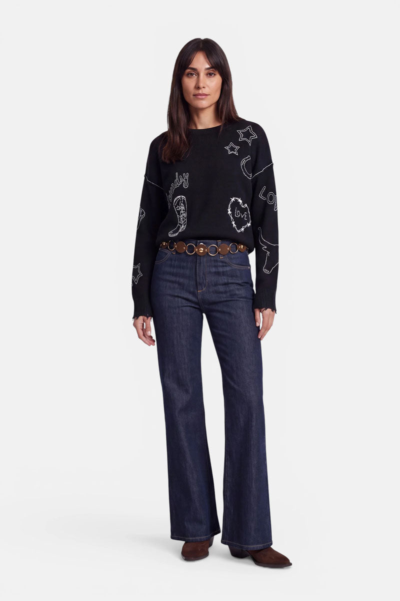 Eine Frau mit langen braunen Haaren trägt einen schwarzen Pullover mit weißen, kritzelartigen Western-Stickereien, dunkelblaue Schlagjeans, einen braunen Gürtel mit runden Verzierungen und braune Schuhe. Sie steht mit dem Blick nach vorne vor einem schlichten weißen Hintergrund.