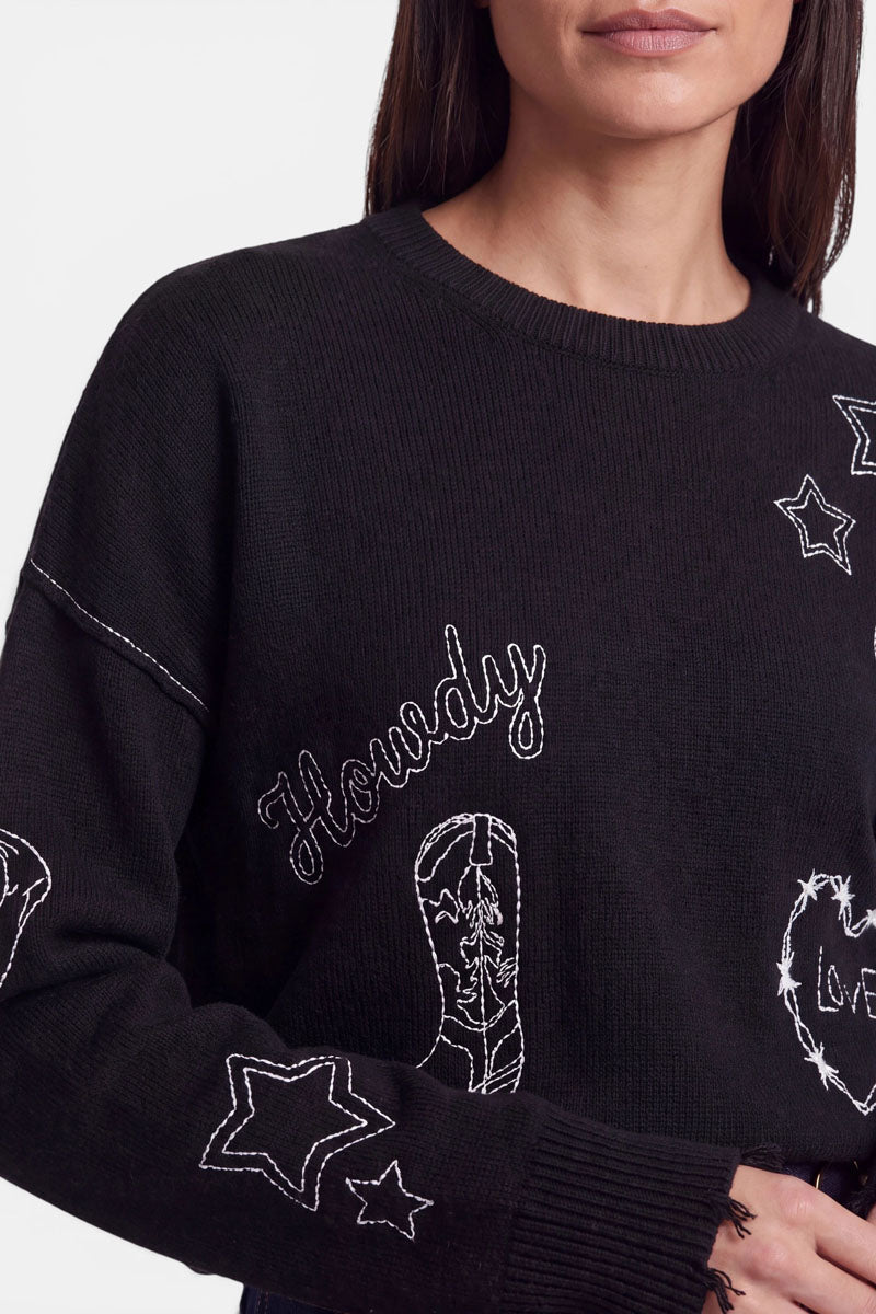 Eine Frau trägt einen schwarzen Pullover aus einer Merino-Kaschmir-Mischung mit weißen Western-Stickereien, darunter Sterne, ein Paar Cowboystiefel, das Wort „Howdy“ und ein Hufeisen. Vor einem schlichten, hellen Hintergrund sind nur ihr unterer Gesichtsbereich und ihr Oberkörper zu sehen.