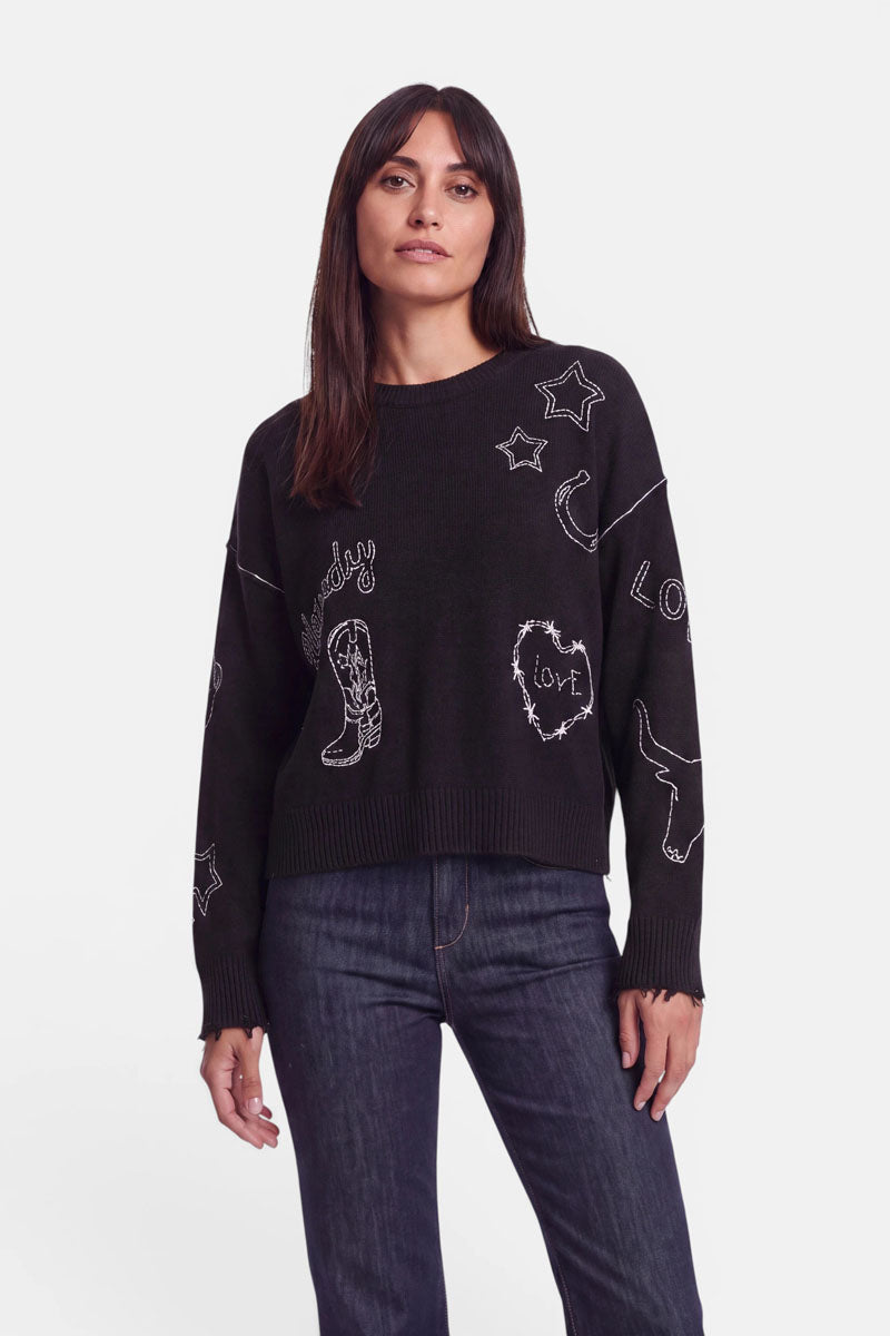 Eine Frau mit langen braunen Haaren trägt einen locker sitzenden Pullover aus einer Merino-Kaschmir-Mischung, dessen schwarzer Stoff mit weißen, im Doodle-Stil gehaltenen Western-Stickereien verziert ist – darunter Sterne, ein Mond, Stiefel, Herzen sowie die Wörter „Love“ und „Cowboy“. Dazu trägt sie eine dunkelblaue Jeans und steht vor einem schlichten weißen Hintergrund.