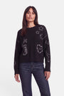 Eine Frau mit langen braunen Haaren trägt einen locker sitzenden Pullover aus einer Merino-Kaschmir-Mischung, dessen schwarzer Stoff mit weißen, im Doodle-Stil gehaltenen Western-Stickereien verziert ist – darunter Sterne, ein Mond, Stiefel, Herzen sowie die Wörter „Love“ und „Cowboy“. Dazu trägt sie eine dunkelblaue Jeans und steht vor einem schlichten weißen Hintergrund.