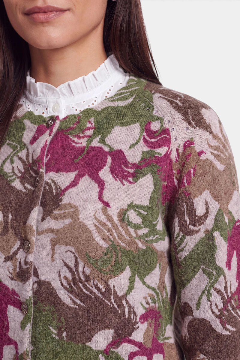 Eine Frau trägt eine weiße Bluse mit Rüschen am Kragen unter einer geknöpften Strickjacke mit einem lebhaften Pferde-Print in Rosa-, Grün-, Braun- und Beigetönen. Die Strickjacke ist aus weicher Alpaka-Wolle gefertigt. Zu sehen sind nur ihr unterer Gesichtsbereich, ihr Hals und ihr Oberkörper. Der Hintergrund ist einfarbig hellgrau.