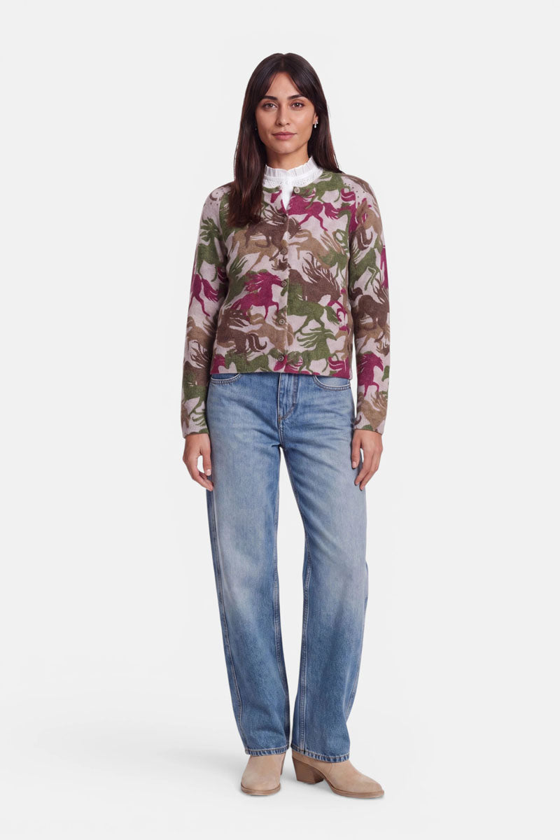 Eine Frau steht mit dem Blick nach vorne und trägt eine Strickjacke mit Camouflage-Muster in Grün, Beige und Rosa aus kuscheliger Alpaka-Wolle über einem weißen Hemd mit Kragen, einer lockeren blauen Jeans und beigen Stiefeln. Sie hat langes braunes Haar und einen ausdruckslosen Gesichtsausdruck und steht vor einem schlichten weißen Hintergrund.