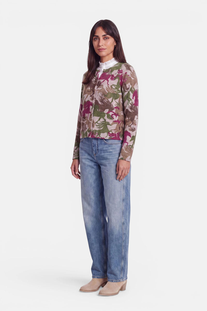 Eine Frau steht mit dem Blick nach vorne. Sie trägt eine gemusterte Strickjacke aus weicher Alpaka-Wolle in Rosa-, Grün- und Beigetönen über einer weißen Bluse mit Rüschenkragen, hellblauen Jeans mit geradem Bein und beigen Schuhen. Sie hat langes dunkles Haar und einen neutralen Gesichtsausdruck. Der Hintergrund ist schlicht weiß.