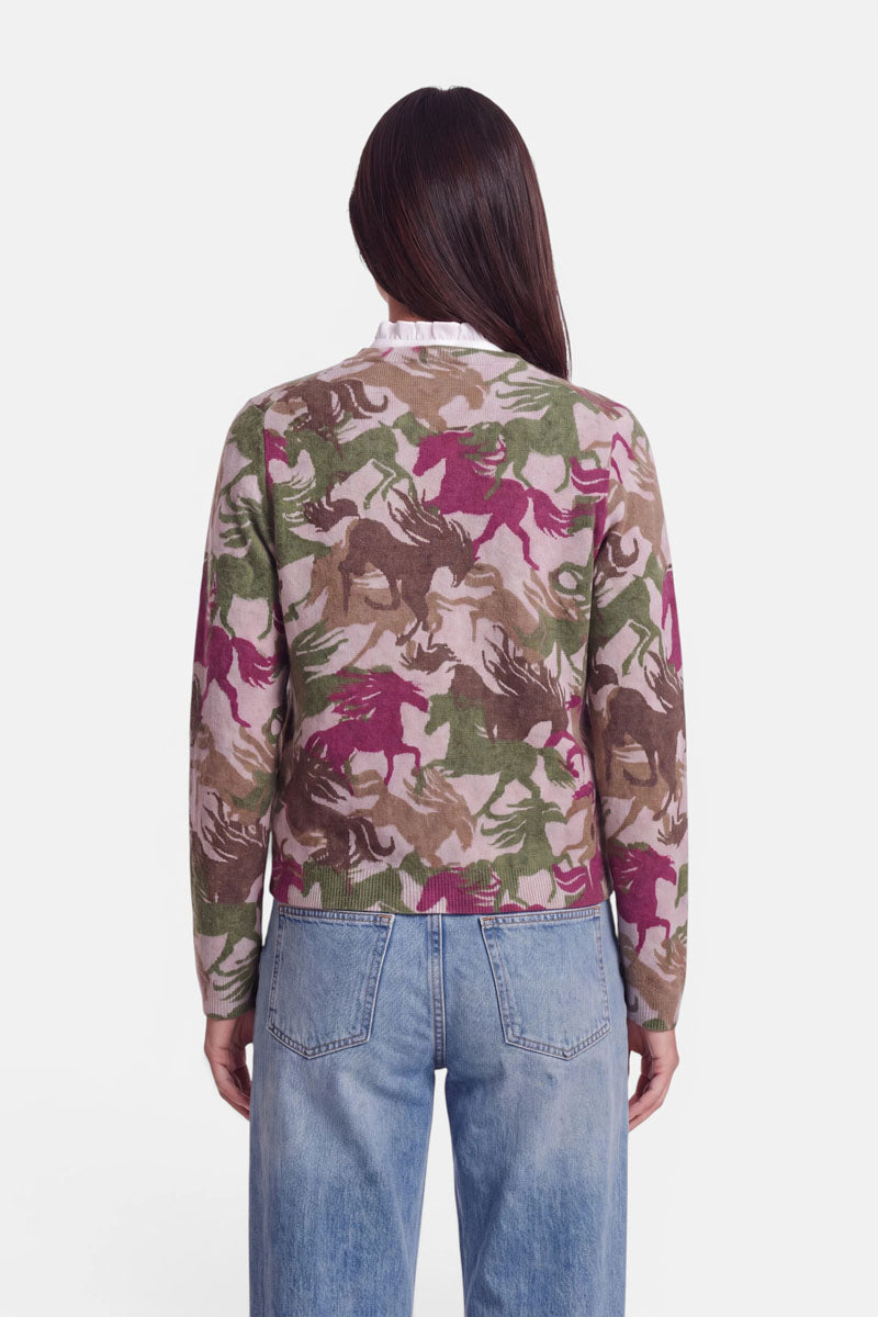 Eine Frau steht mit dem Rücken zur Kamera. Sie trägt eine Strickjacke aus weicher Alpaka-Wolle mit einem camouflageartigen Pferde-Print in Rosa, Grün, Beige und Braun, dazu eine hellblaue Jeans mit hoher Taille. Der Hintergrund ist einfarbig weiß. Ihr dunkles Haar ist glatt und offen.