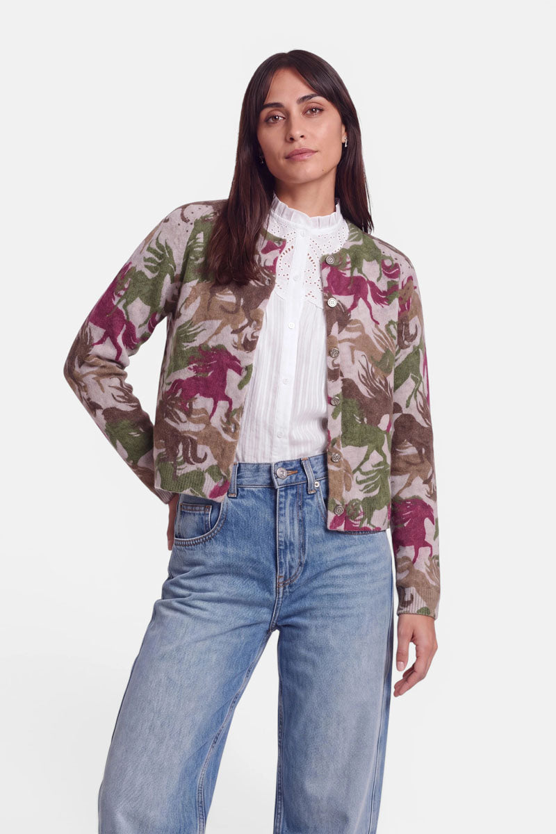 Eine Frau mit glattem braunem Haar, die eine weiße Spitzenbluse, lockere blaue Jeans und eine beige Strickjacke aus Alpaka-Wolle mit grünen, braunen und lilafarbenen Pferdemotiven trägt, steht selbstbewusst mit einer Hand in der Hosentasche vor einem schlichten weißen Hintergrund.