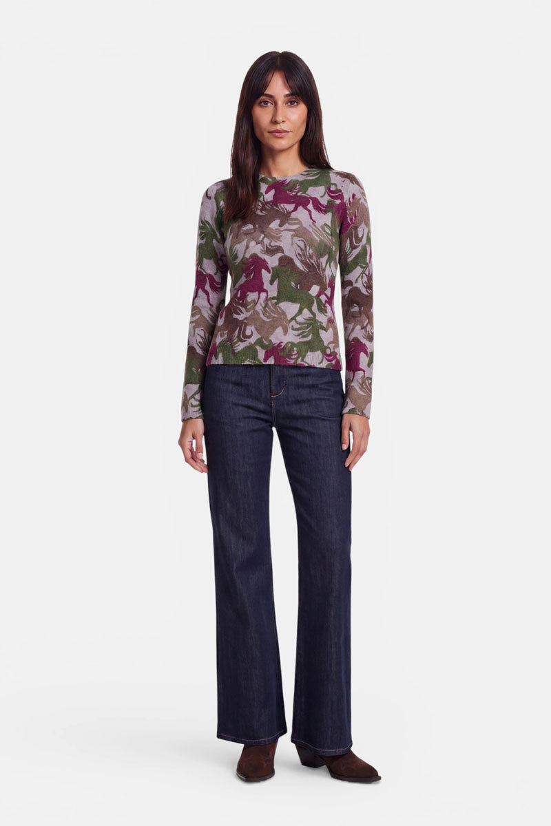 Eine Frau steht mit dem Blick nach vorne und trägt einen eng anliegenden, langärmeligen Pullover aus Alpaka-Wolle mit mehrfarbigem Camouflage-Muster, dazu dunkelblaue Schlagjeans und braune Schuhe. Sie hat glattes braunes Haar, ist natürlich geschminkt und blickt mit neutralem Gesichtsausdruck vor einem weißen Hintergrund in die Kamera.