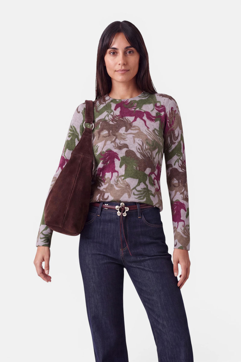 Eine Frau mit glattem, dunklem Haar steht mit dem Blick nach vorne. Sie trägt einen langärmeligen Pullover aus Alpaka-Wolle mit einem bunten Pferdemotiv, dunkelblaue Jeans mit Nietenverzierungen, einen braunen Gürtel mit einer ausgefallenen Schnalle und eine große braune Umhängetasche. Der Hintergrund ist einfarbig weiß.