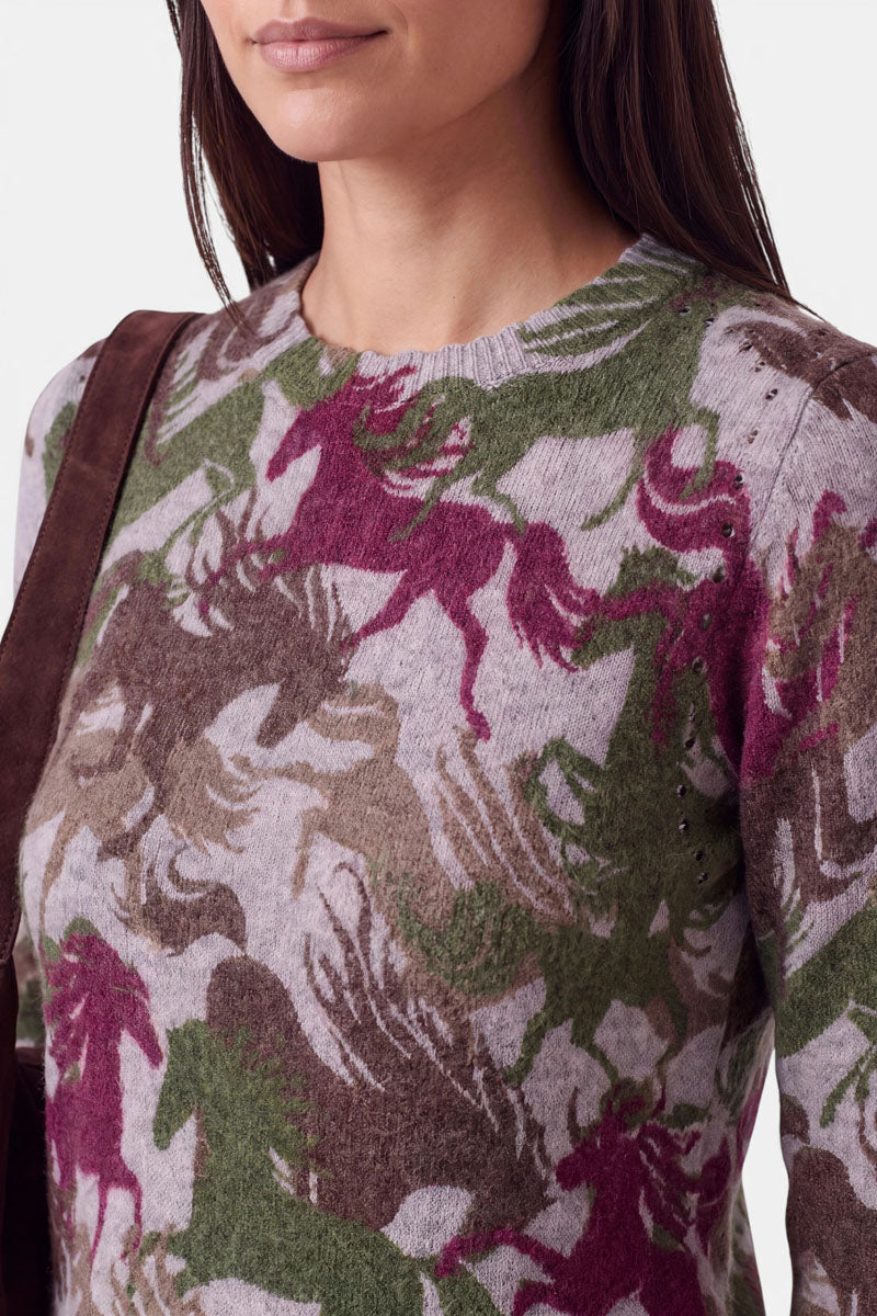 Eine Frau mit glattem braunem Haar trägt einen Pullover aus Alpaka-Wolle mit einem sich überlappenden Muster aus burgunderroten, grünen und braunen Einhörnern. Der Pullover hat einen gerippten Rundhalsausschnitt und dezente Nietendetails, die ihm einen modernen Touch verleihen. Über der Schulter trägt sie einen braunen Riemen, wahrscheinlich von einer Tasche. Zu sehen ist nur ihr Oberkörper.
