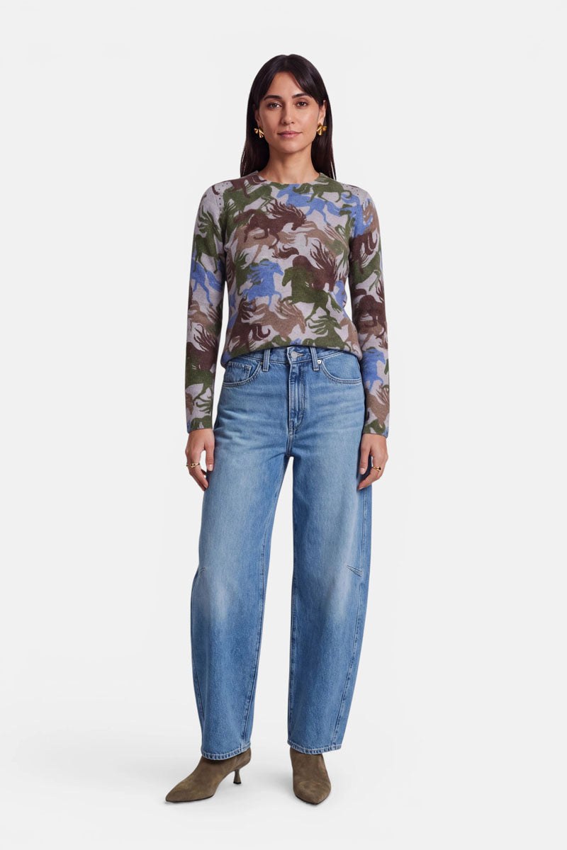 Eine Frau mit langen dunklen Haaren trägt ein eng anliegendes, langärmeliges, mehrfarbiges Oberteil mit Camouflage-Muster und dezenten Nietendetails, hellblaue, locker sitzende Jeans mit hoher Taille sowie braune Stiefel mit spitzer Zehenpartie. Sie steht mit dem Gesicht nach vorne vor einem schlichten weißen Hintergrund.