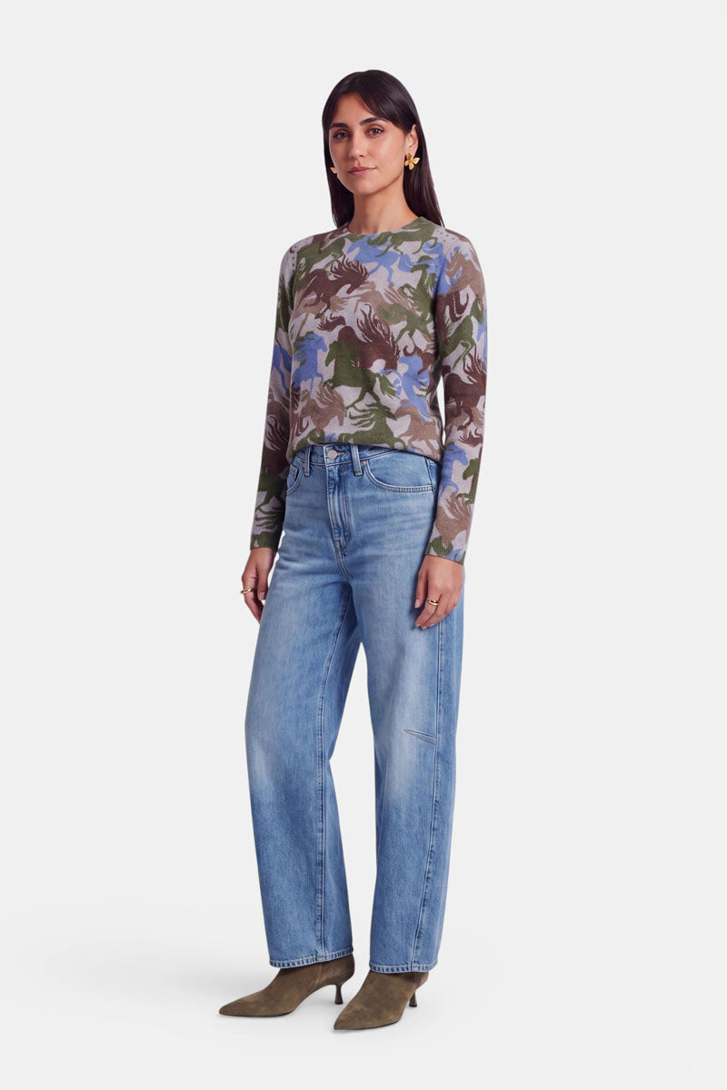 Eine Frau mit glattem, dunklem Haar steht mit dem Blick nach vorne. Sie trägt ein langärmeliges Shirt mit Camouflage-Muster und dezenten Nietendetails, eine locker sitzende hellblaue Jeans, olivgrüne Stiefeletten mit Absatz sowie gelbe Blumen-Ohrringe. Der Hintergrund ist schlicht weiß.