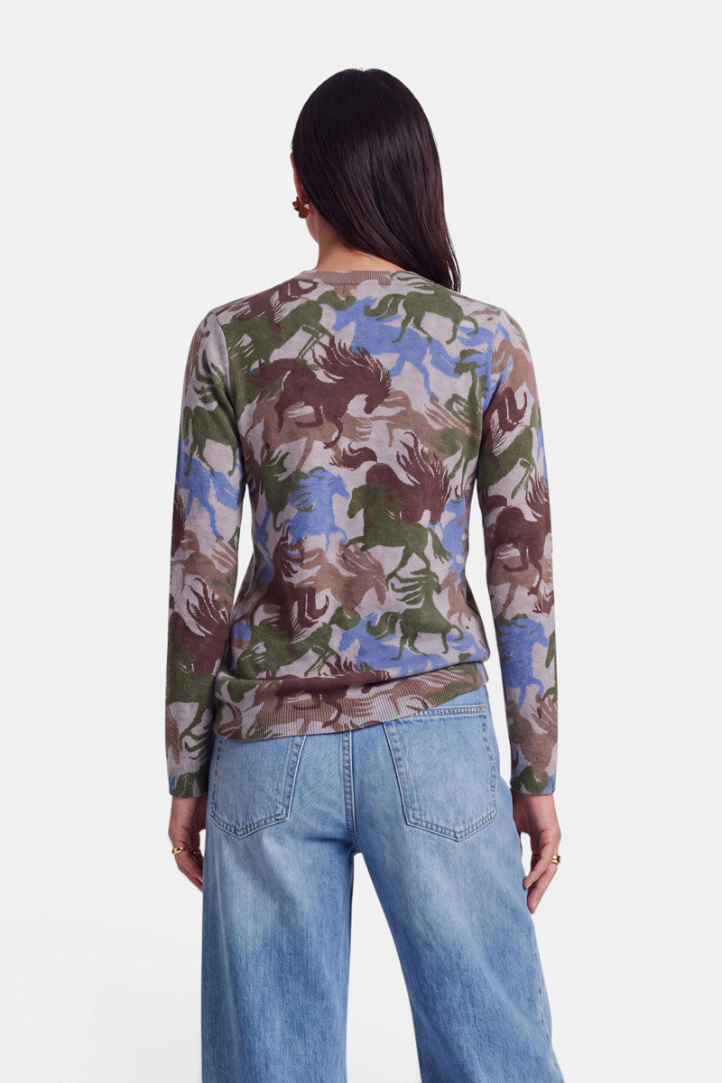 Eine Frau mit langen dunklen Haaren steht mit dem Rücken zur Kamera und trägt einen Pullover aus Alpaka-Wolle mit einem abstrakten Muster in Grün, Braun, Blau und Beige; der Pullover hat lange Ärmel. Außerdem trägt sie eine hellblaue Jeans mit hoher Taille und Nietenverzierungen und steht vor einem schlichten weißen Hintergrund.