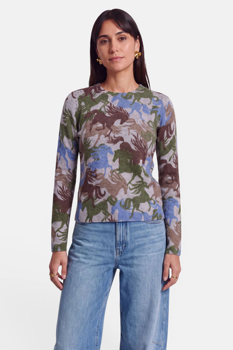 Eine Frau steht vor einem weißen Hintergrund und trägt einen Pullover aus Alpaka-Wolle mit einem abstrakten Pferde-Print in Grün, Braun und Blau sowie hellblaue Jeans. Sie hat langes dunkles Haar, goldene Ohrringe und einen neutralen Gesichtsausdruck; ihre Arme liegen entspannt an den Seiten.