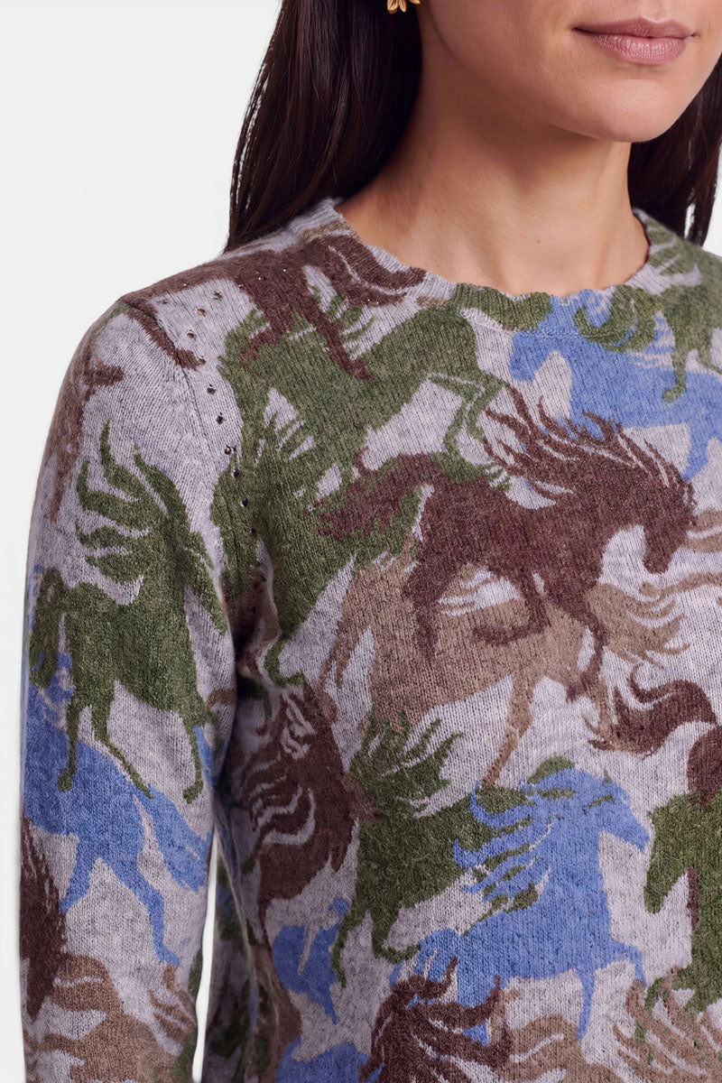 Eine Frau trägt einen Pullover aus Alpaka-Wolle mit einem sich wiederholenden Pferdemuster aus galoppierenden Pferden in Braun, Grün und Blau auf hellgrauem Hintergrund. Zu sehen sind nur ihr Oberkörper, ihre Schulter und der untere Teil ihres Gesichts. Sie hat langes braunes Haar und trägt kleine goldene Ohrringe.