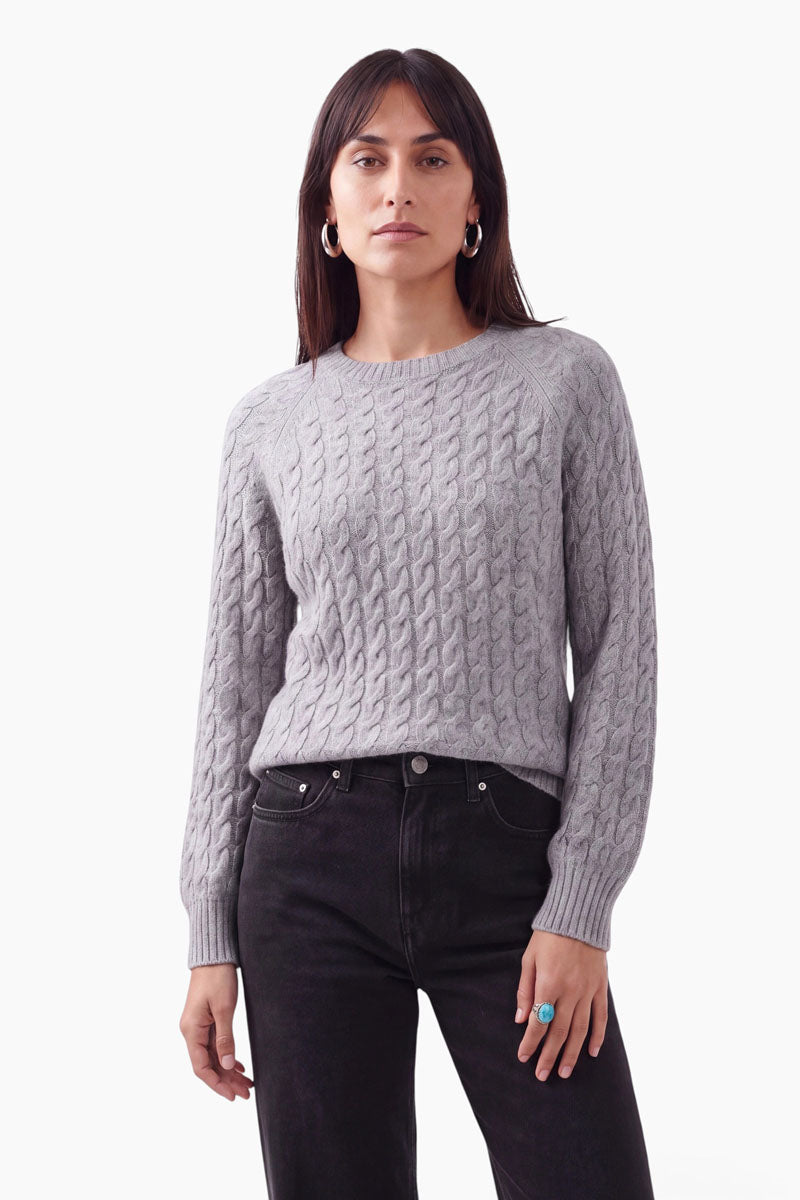 Eine Frau mit langen braunen Haaren trägt einen hellgrauen Zopfstrickpullover mit Merinoanteil und eine schwarze Jeans. Sie steht mit neutralem Gesichtsausdruck, silbernen Creolen und einem türkisfarbenen Ring vor einem schlichten weißen Hintergrund.