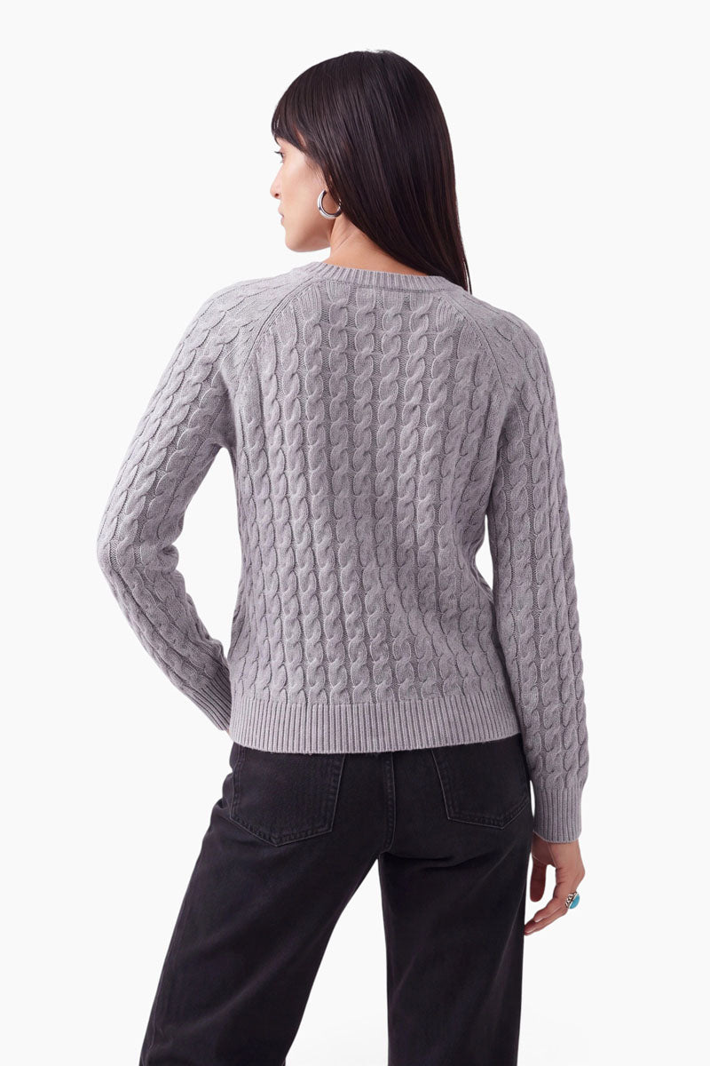 Eine Frau mit glattem, dunklem Haar, von hinten gesehen, trägt einen hellgrauen Zopfstrickpullover mit Merinoanteil und eine schwarze Jeans. Sie hat silberne Creolen und einen türkisfarbenen Ring und steht vor einem schlichten weißen Hintergrund.