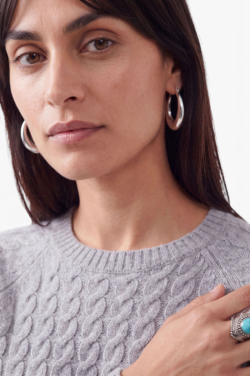 Eine Frau mit glattem, dunklem Haar trägt einen hellgrauen Zopfstrickpullover mit einem hohen Merinoanteil, große silberne Creolen und einen türkisfarbenen Ring. Mit ausdruckslosem Gesicht blickt sie ruhig in die Kamera, während ihre Hand auf der gegenüberliegenden Schulter ruht.