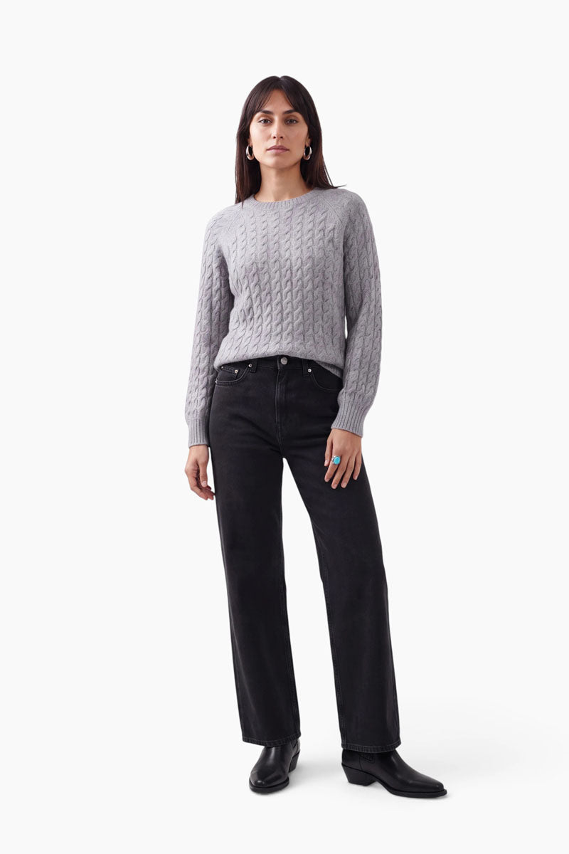 Eine Frau steht vor einem weißen Hintergrund und trägt einen hellgrauen Zopfstrickpullover mit Merinoanteil, eine schwarze A-Linien-Jeans mit hoher Taille und weitem Bein sowie schwarze Stiefeletten. Sie hat langes braunes Haar, Creolen und einen türkisfarbenen Ring, ihren Gesichtsausdruck hält sie neutral und ihre Körperhaltung ist entspannt.