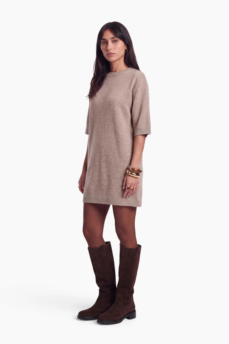 Eine Frau mit langen dunklen Haaren trägt ein beiges Strickkleid mit Merinoanteil und halblangen Ärmeln. Am linken Handgelenk trägt sie goldene Armbänder und große Ringe. Zu dem kurzen Kleid trägt sie kniehohe dunkelbraune Stiefel. Sie steht vor einem schlichten weißen Hintergrund und blickt nach vorne.