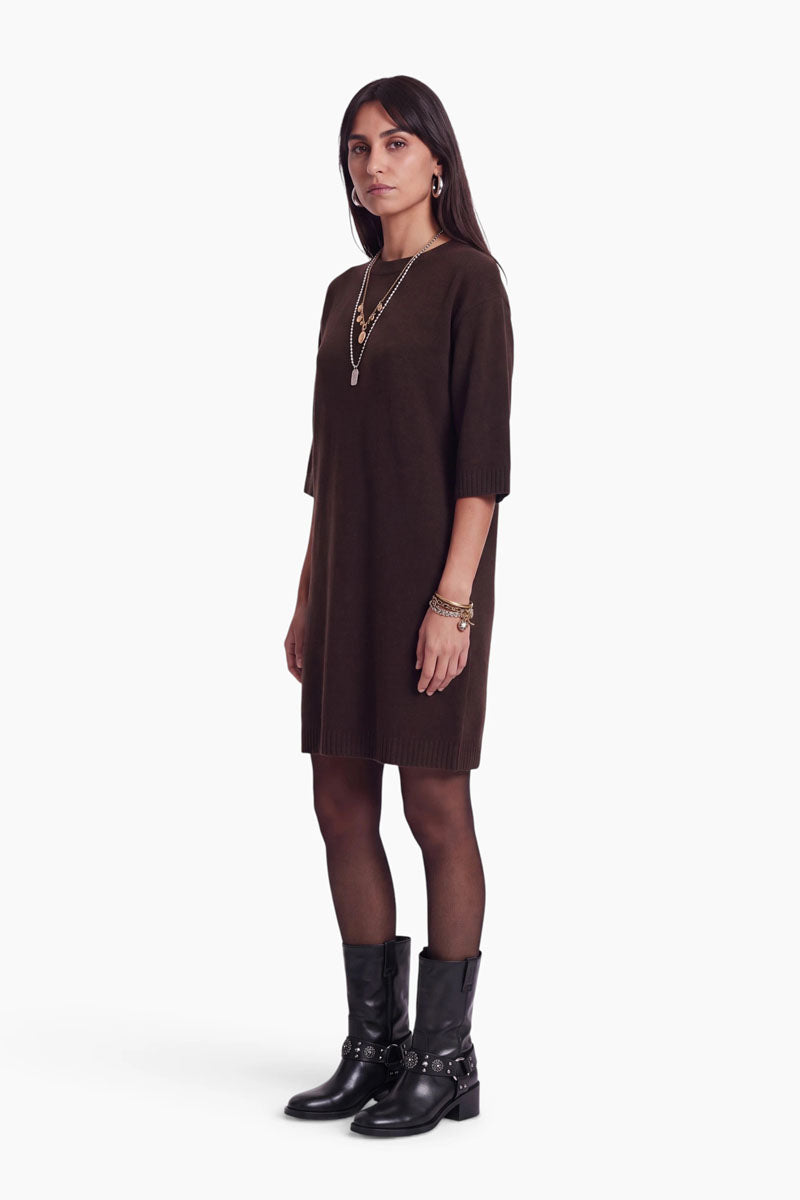 Eine Frau mit glattem, dunklem Haar trägt ein lockeres, dunkelbraunes Strickkleid mit Merinoanteil und halben Ärmeln, eine schwarze Strumpfhose sowie schwarze, wadenhohe Stiefel mit Schnallen. Als Accessoires trägt sie mehrreihige Halsketten, Armbänder und Creolen und steht in einer neutralen Pose vor einem weißen Hintergrund.
