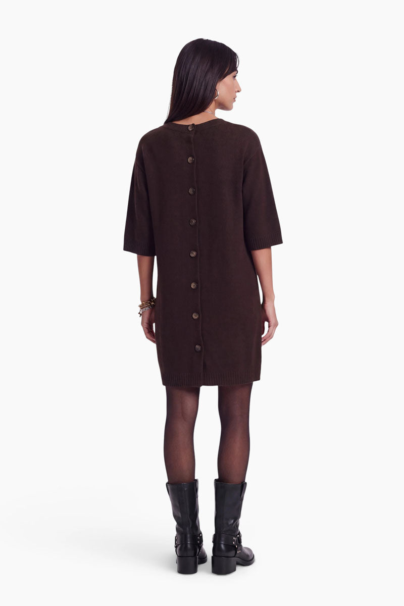 Eine Frau steht mit dem Rücken zur Kamera und trägt ein dunkles, knielanges Strickkleid mit Merinoanteil und einer Knopfreihe am Rücken. Das kurze Kleid hat halblange Ärmel und passt perfekt zu ihrer dunklen, glatten Frisur. Sie trägt schwarze Stiefeletten mit Absatz, transparente schwarze Strumpfhosen sowie Armbänder und Ohrringe als Accessoires.