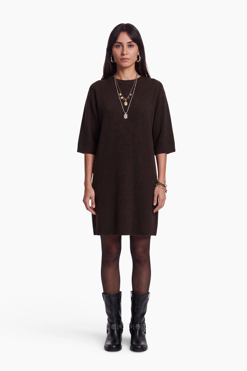 Eine Frau steht mit dem Blick nach vorne und trägt ein lockeres, dunkelbraunes, kurzes Kleid mit ellbogenlangen Ärmeln, eine schwarze Strumpfhose und schwarze Stiefel mit silbernen Schnallen. Ihr glattes braunes Haar wird durch goldene Halsketten, Creolen und Armbänder ergänzt. Der Hintergrund ist weiß.