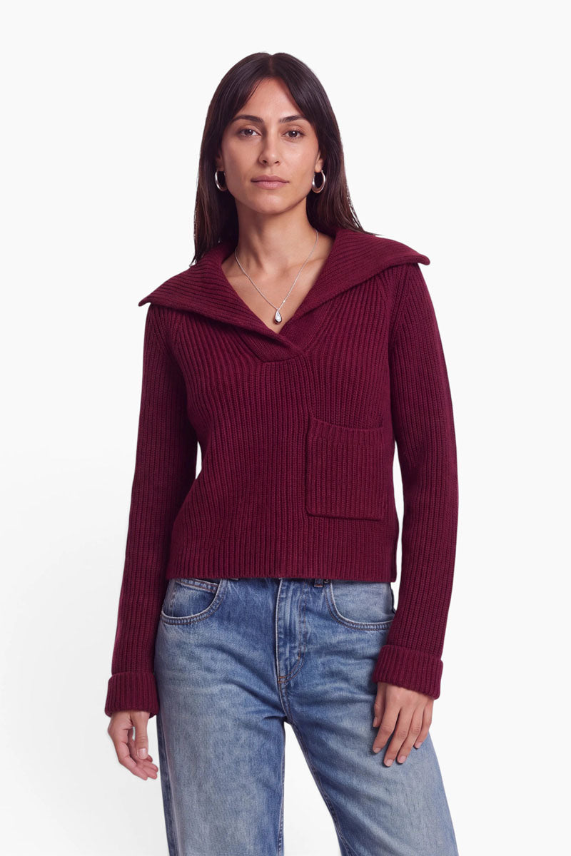 Eine Frau mit glattem, dunklem Haar trägt einen gerippten, weinroten Strickpullover mit V-Ausschnitt und weitem Kragen aus einer edlen Woll-Kaschmir-Mischung, dazu eine Brusttasche, kombiniert mit hellblauen Jeans. Sie steht entspannt frontal mit locker hängenden Armen, trägt eine Halskette mit Anhänger und Creolen-Ohrringe vor einem schlichten weißen Hintergrund.