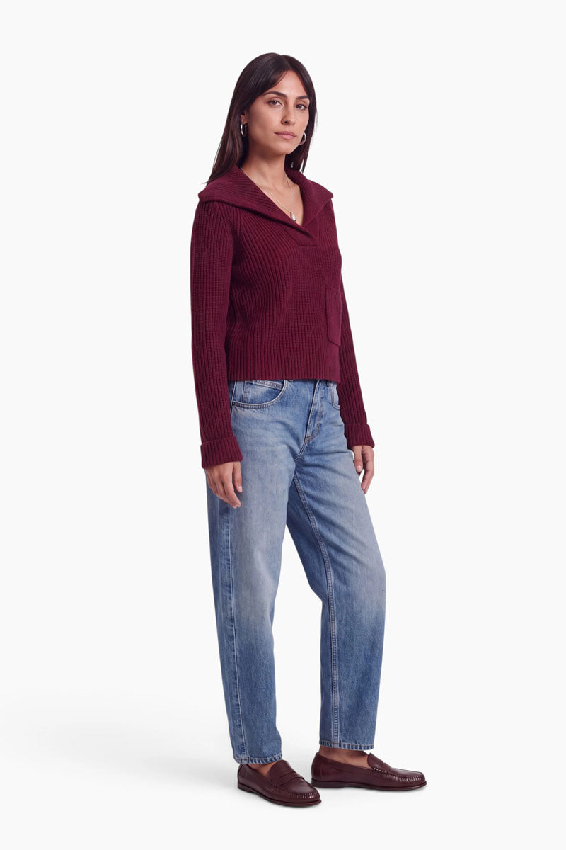 Eine Frau mit langen dunklen Haaren trägt einen burgunderroten Strickpullover mit Kragen und Brusttasche, locker sitzende Bluejeans und braune Slipper. Der Pullover besteht aus einer weichen Woll-Kaschmir-Mischung, die für zusätzlichen Tragekomfort sorgt. Sie steht mit leicht nach rechts gewandtem Blick vor einem weißen Hintergrund und hat einen neutralen Gesichtsausdruck.