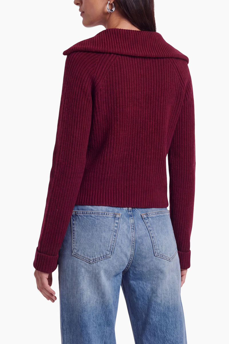 Eine Frau steht mit dem Rücken zur Kamera und trägt einen gerippten Strickpullover aus einem Woll-Kaschmir-Mix in Dunkelrot mit breitem Kragen sowie eine hellblaue, hoch taillierte Jeans mit geradem Bein. Sie hat langes dunkles Haar, trägt sichtbare Creolen und lässt ihren linken Arm entspannt an der Seite hängen, vor einem weißen Hintergrund.