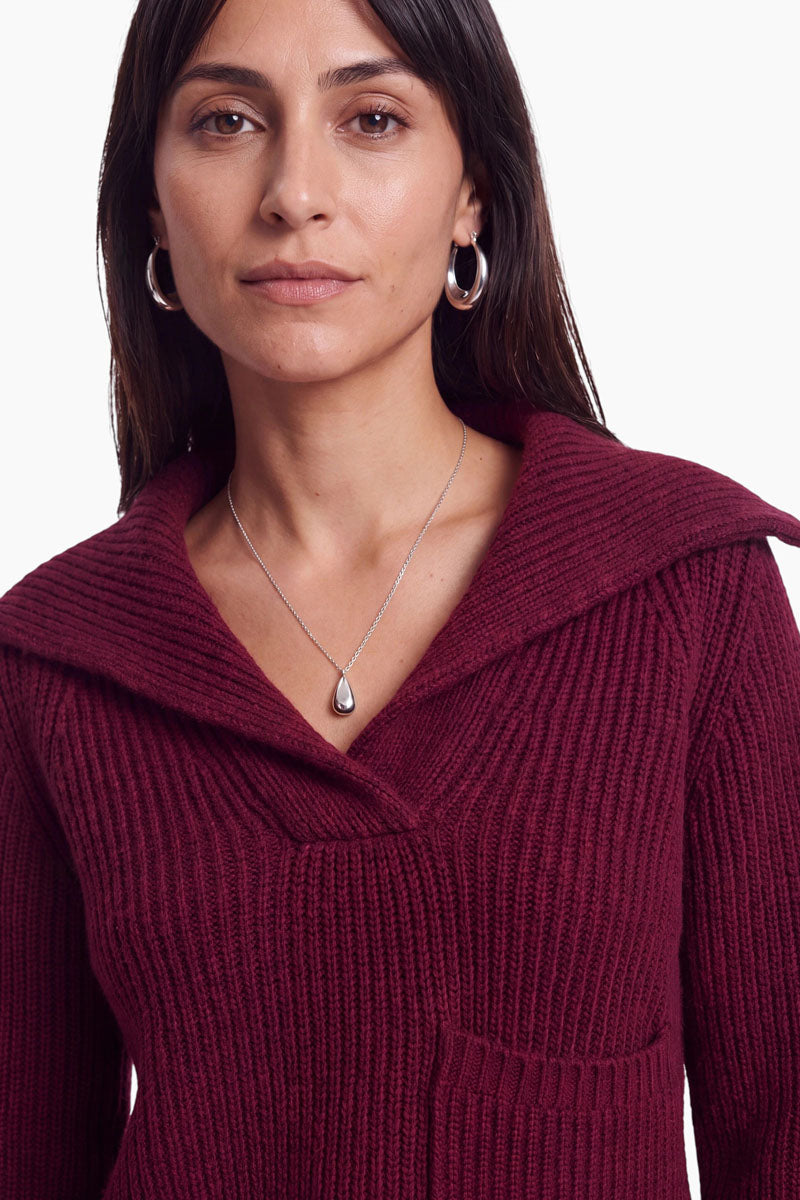 Eine Frau mit langen dunklen Haaren trägt einen gerippten, weinroten Strickpullover aus einer Woll-Kaschmir-Mischung mit breitem Kragen, silberne Creolen und eine Halskette mit einem tropfenförmigen Anhänger. Sie blickt mit ausdruckslosem Gesicht nach vorne vor einem schlichten weißen Hintergrund.