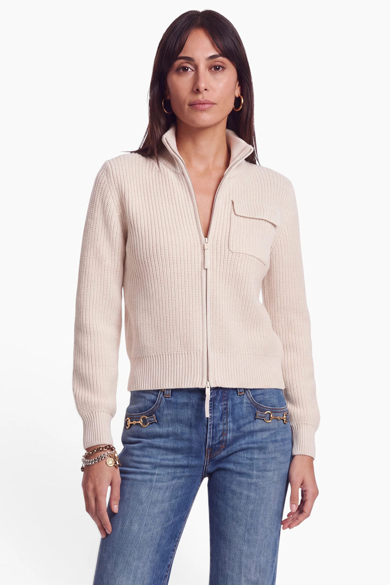 Eine Frau mit glattem, dunklem Haar trägt eine beige Strickjacke aus einer Woll-Kaschmir-Mischung mit einer Brusttasche und einem Zwei-Wege-Reißverschluss, dazu blaue Jeans mit goldenen Akzenten, goldene Creolen und Armbänder; sie steht vor einem schlichten weißen Hintergrund.