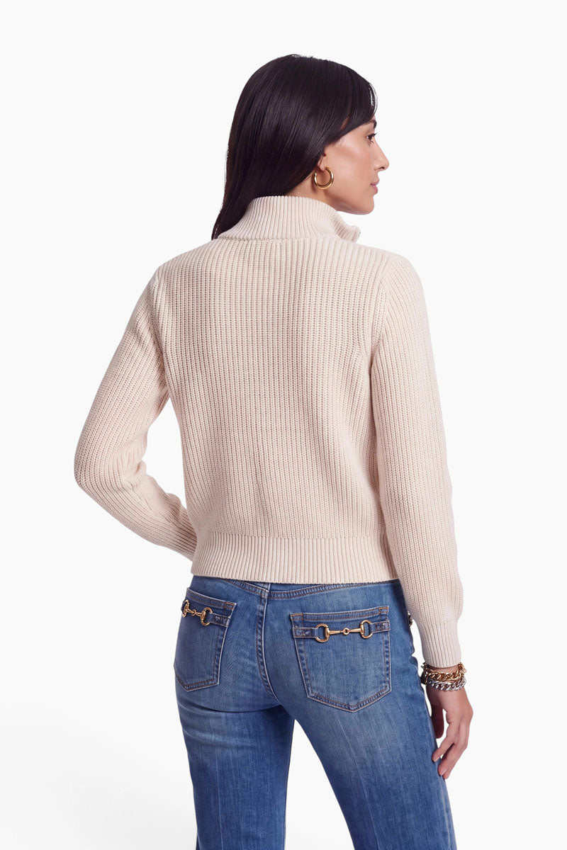 Eine Frau mit langen dunklen Haaren steht mit dem Rücken zur Kamera. Sie trägt einen cremefarbenen, gerippten Rollkragenpullover aus einer luxuriösen Wolle-Kaschmir-Mischung sowie eine blaue Jeans mit goldenen Kettenverzierungen an den Gesäßtaschen. Ihre linke Hand ruht auf ihrer Hüfte, und sie trägt goldene Creolen sowie Armbänder. Der Hintergrund ist schlicht weiß, wodurch ihr gemütliches Strickjacken-Outfit besonders gut zur Geltung kommt.