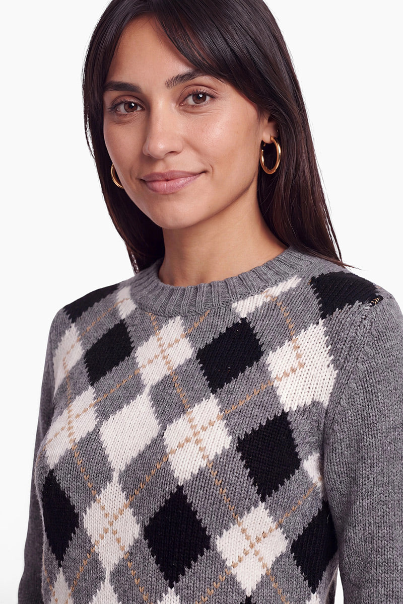 Eine Frau mit langen dunklen Haaren und goldenen Creolen lächelt sanft. Sie trägt einen Strickpullover aus einer Woll-Kaschmir-Mischung mit einem schwarz-weiß-beigen Argyle-Muster. Der Hintergrund ist schlicht weiß.