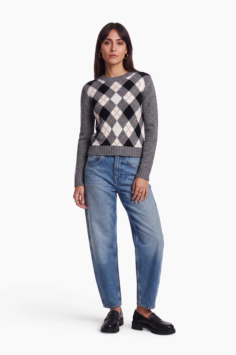Eine Frau steht mit dem Blick nach vorne gerichtet da. Sie trägt einen grauen Streickpullover mit Argyle-Muster aus einer Woll-Kaschmir-Mischung, eine lockere blaue Jeans und schwarze Slipper. Sie hat glattes, dunkles Haar, goldene Creolen und einen ausdruckslosen Gesichtsausdruck; eine Hand steckt in ihrer Hosentasche. Der Hintergrund ist schlicht weiß.
